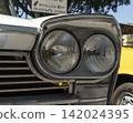 Tavernes Blanques, Spain - June 14, 2026. Close up shot of twin front headlight assembly and chrome trim on a vintage white Seat car parked under a tree in a city setting 142024395