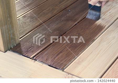 Brown oil for protection of wood by staining. Painting a larch for protecting and pests. Woodworker applying finish to terrace deck board. Repairman varnishing with brush wooden plank floor surface. Brown oil for protection of wood by staining. Painting a larch for protecting and pests. Woodworker applying finish to terrace deck board. Repairman varnishing with brush wooden plank floor surface. 142024440