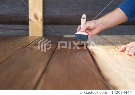 Brown oil for protection of wood by staining. Painting a larch for protecting and pests. Woodworker applying finish to terrace deck board. Repairman varnishing with brush wooden plank floor surface. Brown oil for protection of wood by staining. Painting a larch for protecting and pests. Woodworker applying finish to terrace deck board. Repairman varnishing with brush wooden plank floor surface. 142024444