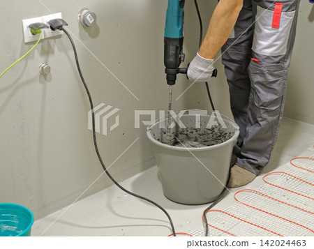 Construction worker preparing tile surface, mixing mortar substrate for flooring, underfloor heating mat. Workman combines adhesive in renovation setting. Operating power tool during bathroom upgrade Construction worker preparing tile surface, mixing mortar substrate for flooring, underfloor heating mat. Workman combines adhesive in renovation setting. Operating power tool during bathroom upgrade 142024463