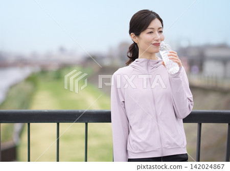 一位中年婦女邊走邊喝瓶裝水。 一位中年婦女邊走邊喝瓶裝水。 142024867