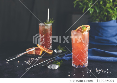 Pink Grapefruit Mezcal Paloma Cocktail in highball glass with water drops on table in real home. New life concept of quarantine, shallow depth of the field, dark background. Pink Grapefruit Mezcal Paloma Cocktail in highball glass with water drops on table in real home. New life concept of quarantine, shallow depth of the field, dark background. 142026005