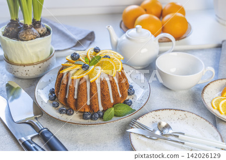 Orange bundt cake with blueberry surrounded fruits, plant and cutlery on light table near window. Family breakfast concept Orange bundt cake with blueberry surrounded fruits, plant and cutlery on light table near window. Family breakfast concept 142026129