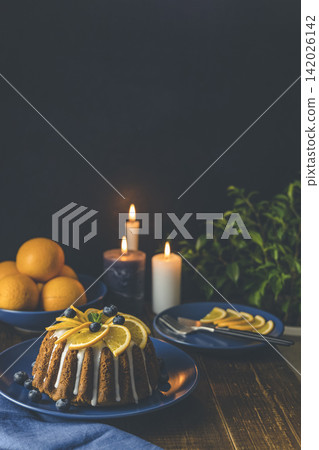 Orange bundt cake with blueberry surrounded fruits, candles, plant and cutleries on dark wooden background. Orange bundt cake with blueberry surrounded fruits, candles, plant and cutleries on dark wooden background. 142026142