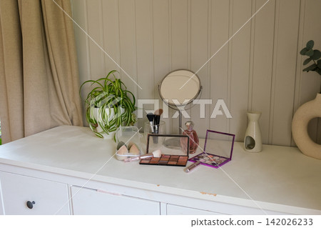 Vanity round mirror and make up items arranged on desk under lamp, palettes and brushes, open eyeshadow, cotton pads. Makeup and self care at home concept. Personal beauty products items on table. Vanity round mirror and make up items arranged on desk under lamp, palettes and brushes, open eyeshadow, cotton pads. Makeup and self care at home concept. Personal beauty products items on table. 142026233
