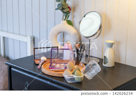 Vanity round mirror and make up items arranged on desk under lamp, palettes and brushes, open eyeshadow, cotton pads. Makeup and self care at home concept. Personal beauty products items on table. Vanity round mirror and make up items arranged on desk under lamp, palettes and brushes, open eyeshadow, cotton pads. Makeup and self care at home concept. Personal beauty products items on table. 142026234