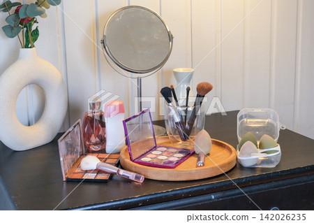 Vanity round mirror and make up items arranged on desk under lamp, palettes and brushes, open eyeshadow, cotton pads. Makeup and self care at home concept. Personal beauty products items on table. Vanity round mirror and make up items arranged on desk under lamp, palettes and brushes, open eyeshadow, cotton pads. Makeup and self care at home concept. Personal beauty products items on table. 142026235