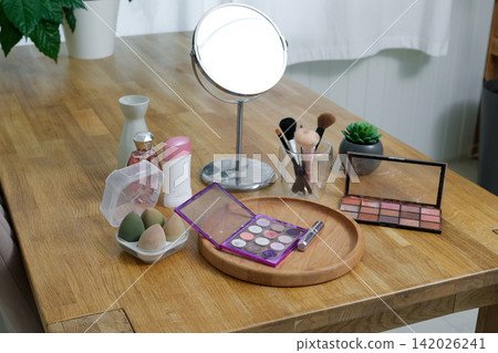 Vanity round mirror and make up items arranged on desk under lamp, palettes and brushes, open eyeshadow, cotton pads. Makeup and self care at home concept. Personal beauty products items on table. Vanity round mirror and make up items arranged on desk under lamp, palettes and brushes, open eyeshadow, cotton pads. Makeup and self care at home concept. Personal beauty products items on table. 142026241