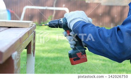 Worker man working with Screwdriver. Person using a red cordless drill with a metal spiral drill bit in a workshop, construction, diy project task. Accu battery hand drill. Screwing a screw assembling Worker man working with Screwdriver. Person using a red cordless drill with a metal spiral drill bit in a workshop, construction, diy project task. Accu battery hand drill. Screwing a screw assembling 142026750