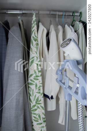 A steam steamer is focused on the background of a wardrobe with clothes on hangers, its metal nozzle and mounting to the rack are visible in detail. Concept: the technology of making clothes look A steam steamer is focused on the background of a wardrobe with clothes on hangers, its metal nozzle and mounting to the rack are visible in detail. Concept: the technology of making clothes look 142027800