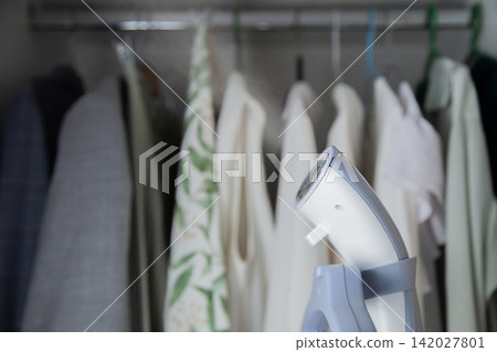 Against the blurred background of a wardrobe with clothes in white and green tones, a white steam steamer placed on the counter is in focus. Concept: household care of things, wardrobe preparation Against the blurred background of a wardrobe with clothes in white and green tones, a white steam steamer placed on the counter is in focus. Concept: household care of things, wardrobe preparation 142027801