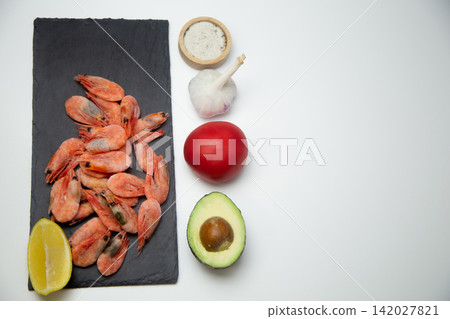 There is a handful of frozen shrimp with lemon on a black board on a white background, and a salt shaker, garlic, tomato, and avocado are lined up next to it. The concept: a neat layout of ingredients There is a handful of frozen shrimp with lemon on a black board on a white background, and a salt shaker, garlic, tomato, and avocado are lined up next to it. The concept: a neat layout of ingredients 142027821