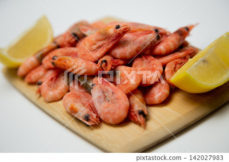 Close-up of frozen pink shrimp on a light wooden board surrounded by two bright lemon slices on a white background. Concept: bright color contrast, gastronomic aesthetics of fresh seafood. High Close-up of frozen pink shrimp on a light wooden board surrounded by two bright lemon slices on a white background. Concept: bright color contrast, gastronomic aesthetics of fresh seafood. High 142027983