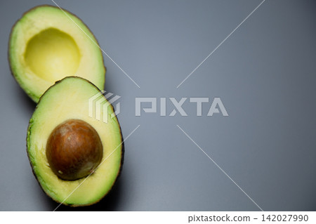 On a gray background, two avocado halves are located on the left side of the frame, the lower one with a stone, the upper one without it, creating a vertical composition. Concept: minimalistic 142027990