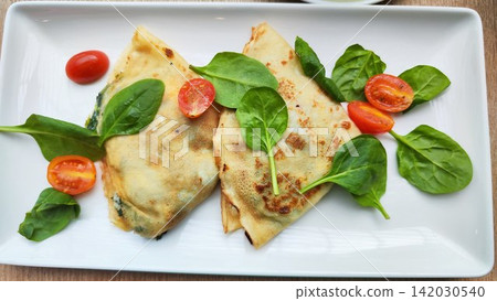 Two folded pancakes, crepes on a rectangular white plate are garnished with fresh spinach leaves (Spinacia oleracea) and halved cherry tomatoes (Solanum lycopersicum var. cerasiforme) Two folded pancakes, crepes on a rectangular white plate are garnished with fresh spinach leaves (Spinacia oleracea) and halved cherry tomatoes (Solanum lycopersicum var. cerasiforme) 142030540