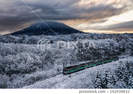 冬日美景徐徐展開:一列當地火車沿著被白雪覆蓋的樹木環繞的鐵軌行駛,遠處是雄偉的雪山羊蹄山。 冬日美景徐徐展開:一列當地火車沿著被白雪覆蓋的樹木環繞的鐵軌行駛,遠處是雄偉的雪山羊蹄山。 142030632