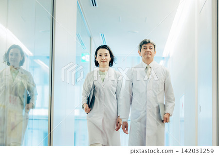 Two researchers in their 50s, wearing white lab coats, walk through a research facility. Two researchers in their 50s, wearing white lab coats, walk through a research facility. 142031259