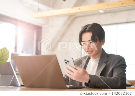 一位商人一邊用電腦工作,一邊查看智慧型手機。 一位商人一邊用電腦工作,一邊查看智慧型手機。 142032102