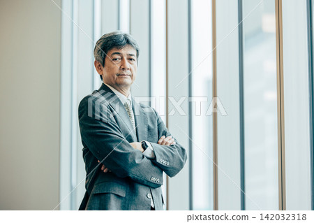 一位資深商人雙手交叉放在胸前,坐在辦公室的窗邊。 一位資深商人雙手交叉放在胸前,坐在辦公室的窗邊。 142032318
