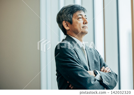 A senior businessman with his arms crossed, sitting by the office window. A senior businessman with his arms crossed, sitting by the office window. 142032320