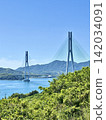 The Tatara Bridge and the blue sea viewed from atop a hill with lush greenery (Shimanami Kaido, Omishima Island) 142034091