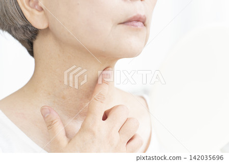 一位老婦人坐在客廳裡，拿著一面小鏡子，擔憂地看著臉上的衰老跡象。 142035696
