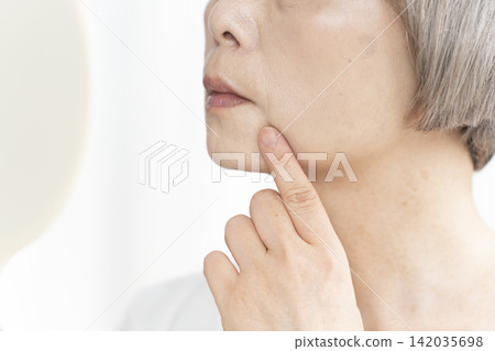 一位老婦人坐在客廳裡，拿著一面小鏡子，擔憂地看著臉上的衰老跡象。 142035698