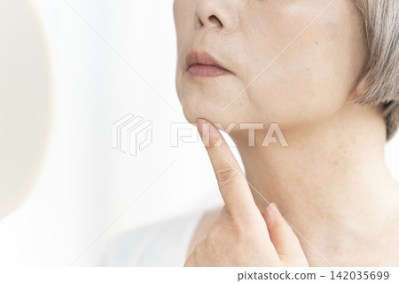 A senior woman in her living room, holding a hand mirror, is concerned about the signs of aging on her face. 142035699