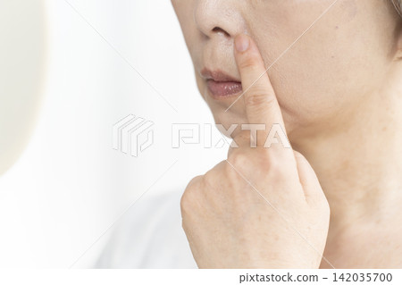 A senior woman in her living room, holding a hand mirror, is concerned about the signs of aging on her face. A senior woman in her living room, holding a hand mirror, is concerned about the signs of aging on her face. 142035700