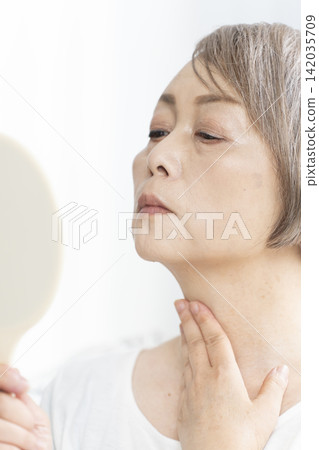 一位老婦人坐在客廳裡,拿著一面小鏡子,擔憂地看著臉上的衰老跡象。 一位老婦人坐在客廳裡,拿著一面小鏡子,擔憂地看著臉上的衰老跡象。 142035709