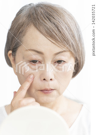 一位老婦人坐在客廳裡,拿著一面小鏡子,擔憂地看著臉上的衰老跡象。 一位老婦人坐在客廳裡,拿著一面小鏡子,擔憂地看著臉上的衰老跡象。 142035721
