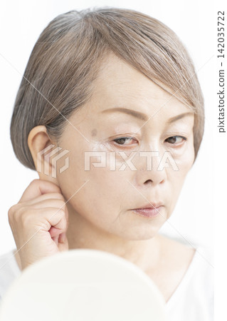 A senior woman in her living room, holding a hand mirror, is concerned about the signs of aging on her face. A senior woman in her living room, holding a hand mirror, is concerned about the signs of aging on her face. 142035722