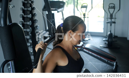 Young woman in black sportswear performing a behind-the-neck lat pulldown exercise on a cable machine in a gym. Back muscle training concept Young woman in black sportswear performing a behind-the-neck lat pulldown exercise on a cable machine in a gym. Back muscle training concept 142036431