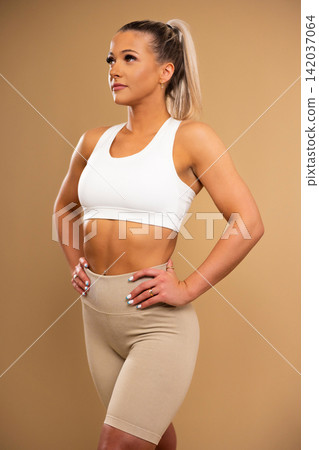 Fit young woman in white sports bra and beige leggings posing confidently against beige background Fit young woman in white sports bra and beige leggings posing confidently against beige background 142037064