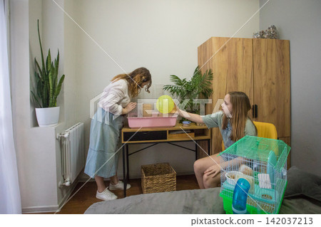 Two people are cleaning a hamster cage. One person is sitting on a bed while the other Two people are cleaning a hamster cage. One person is sitting on a bed while the other 142037213