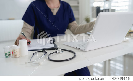 Asian female doctor writing prescription notes while using laptop computer for telemedicine consultation in modern clinic office Asian female doctor writing prescription notes while using laptop computer for telemedicine consultation in modern clinic office 142039446