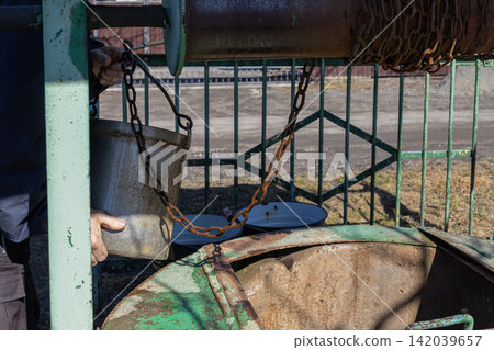 A person holds a metal bucket at an old well using a rusty chain to draw water from the underground source, representing rural life, self-sufficiency and traditional resource usage A person holds a metal bucket at an old well using a rusty chain to draw water from the underground source, representing rural life, self-sufficiency and traditional resource usage 142039657