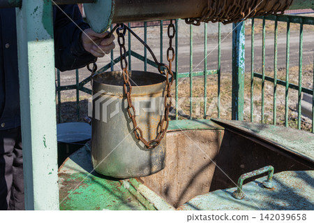 A hand pulls an old metal bucket from a rustic water well using a heavy iron chain mechanism, evoking traditional rural life and the simple act of manual resource collection daily A hand pulls an old metal bucket from a rustic water well using a heavy iron chain mechanism, evoking traditional rural life and the simple act of manual resource collection daily 142039658