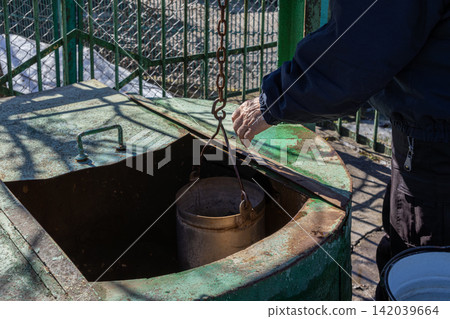 A hand lowers a metal bucket into an old rural water well via a rusty chain mechanism, representing traditional manual labor, village life, and the essential resource of hydration A hand lowers a metal bucket into an old rural water well via a rusty chain mechanism, representing traditional manual labor, village life, and the essential resource of hydration 142039664