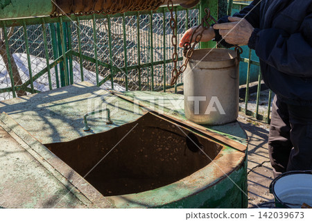 A senior person operates a metal bucket on a chain at an old rural water well, capturing a scene of manual labor and traditional life in a rustic backyard during a bright sunny day A senior person operates a metal bucket on a chain at an old rural water well, capturing a scene of manual labor and traditional life in a rustic backyard during a bright sunny day 142039673