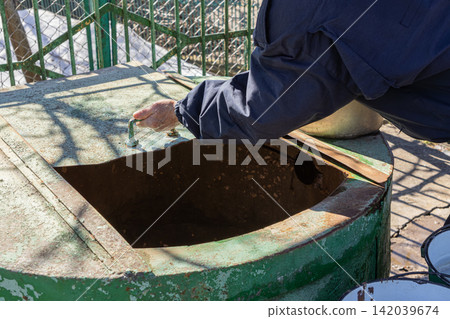 A hand in a blue uniform sleeve opens a rusty green metal cover on an old water well, representing manual labor and infrastructure maintenance tasks in a rural outdoor setting daily 142039674