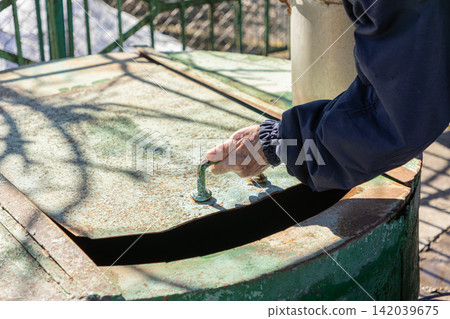 A hand opens the heavy metal lid of a rustic water well, highlighting manual labor and maintenance in a garden setting, emphasizing utility and access to essential natural resources A hand opens the heavy metal lid of a rustic water well, highlighting manual labor and maintenance in a garden setting, emphasizing utility and access to essential natural resources 142039675