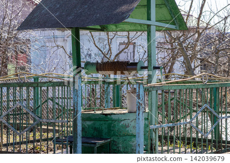 A vintage green water well with a rusty chain and metal bucket stands in a garden surrounded by a metal fence, evoking rural nostalgia and the traditional life of the countryside A vintage green water well with a rusty chain and metal bucket stands in a garden surrounded by a metal fence, evoking rural nostalgia and the traditional life of the countryside 142039679