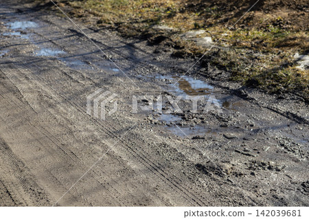 A muddy country road featuring deep water puddles and tire tracks in early spring, illustrating rural terrain, challenging driving conditions, and the damp aftermath of seasonal thaw A muddy country road featuring deep water puddles and tire tracks in early spring, illustrating rural terrain, challenging driving conditions, and the damp aftermath of seasonal thaw 142039681