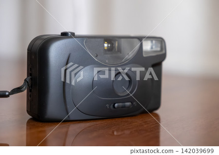 A black vintage compact film camera rests on a polished wooden surface, evoking nostalgia for traditional analog photography and the simple joy of capturing spontaneous memories daily A black vintage compact film camera rests on a polished wooden surface, evoking nostalgia for traditional analog photography and the simple joy of capturing spontaneous memories daily 142039699