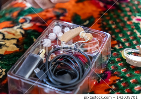 A beige rechargeable hearing aid device lies inside a plastic storage tray with a charging cable and silicone tips to improve audio clarity for individuals with sensory loss issues A beige rechargeable hearing aid device lies inside a plastic storage tray with a charging cable and silicone tips to improve audio clarity for individuals with sensory loss issues 142039701