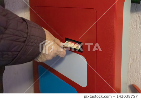 A hand inserts paper currency into a bright red payment kiosk, representing a convenient cash transaction in a public terminal used for fast financial service at a local indoor shop A hand inserts paper currency into a bright red payment kiosk, representing a convenient cash transaction in a public terminal used for fast financial service at a local indoor shop 142039757