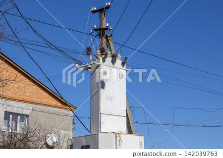 A concrete utility pole with a large white electrical transformer box stands against a clear blue sky, illustrating infrastructure and power distribution in a quiet residential area A concrete utility pole with a large white electrical transformer box stands against a clear blue sky, illustrating infrastructure and power distribution in a quiet residential area 142039763