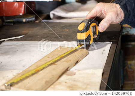 A carpenter holds a yellow measuring tape over a wooden board on a workbench to capture precise dimensions for home renovation projects, representing accuracy in manual labor craft 142039768
