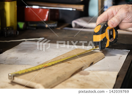 A senior hand uses a yellow metal tape measure to check a pine plank, highlighting precision and measurement in a professional carpentry workshop for home improvement projects A senior hand uses a yellow metal tape measure to check a pine plank, highlighting precision and measurement in a professional carpentry workshop for home improvement projects 142039769